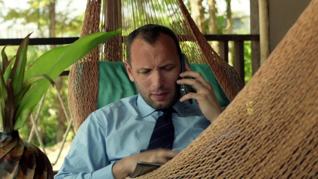 Angry businessman with tablet computer talking on cellphone lying on hammock
