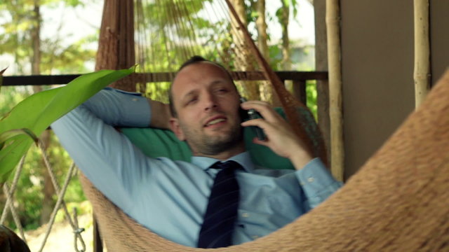 Young businessman talking on cellphone lying on hammock 
