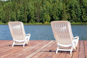 Two white deck chairs on the pier in front of the river in the summer forest