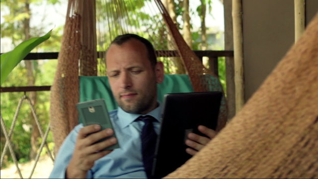 Young businessman comparing data on smartphone and tablet computer on hammock
