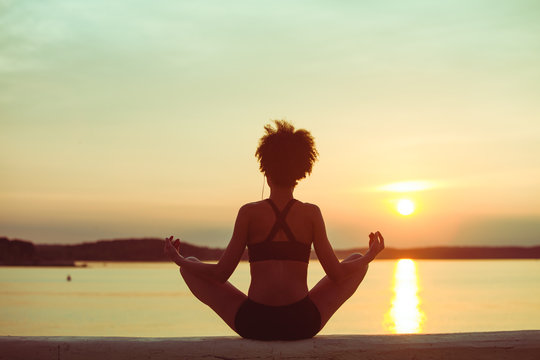 Relaxed fitness attractive young African girl resting after exercising and watching the sun on summer sunset at beach.