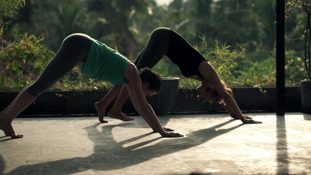 Two young girlfriends on terrace exercising yoga, doing sun salutation pose 
