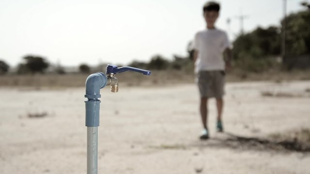 Water shortage concept.Close up Asian boy open metal blue water faucet on hot and dry empty land.