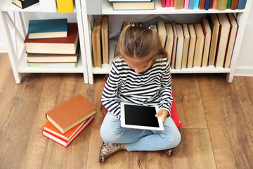Beautiful little girl with tablet and books sitting on floor in library
