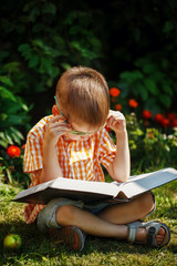 Beautiful kid boy with glasses, reading a book in garden, sitting on grass.