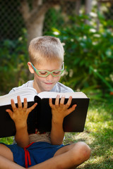 Cute kid boy with glasses, reading a book in garden, sitting on grass.