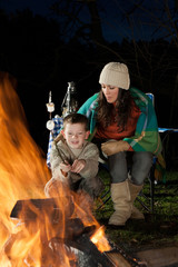 Little boy is sitting with his mother at a bonfire whilst roasting marshmallows.