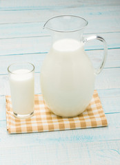 A glass of milk and a milk jug on plaid tablecloth.