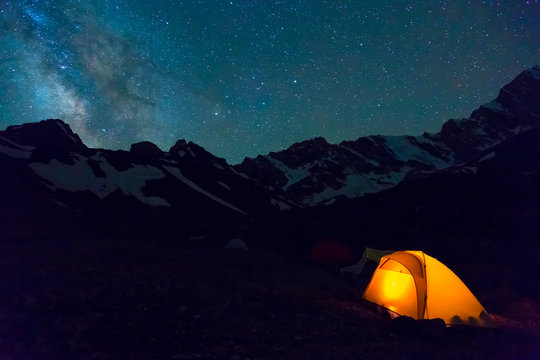 Night mountain landscape with illuminated tent Silhouettes of snowy mountain peaks and edges night sky with many stars and milky way on background illuminated orange tent on foreground