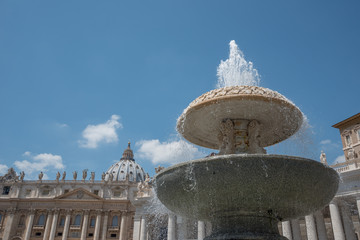 Wasserspeiender Brunnen auf dem Petersplatz in Rom mit blauem Himmel und Teilen des Petersdoms im Hintergrund