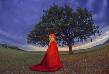 Beautiful blonde woman in old-fashioned dress and red cloak sitting under an oak