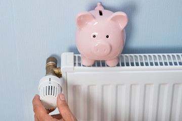Man Holding Thermostat With Piggy Bank On Radiator