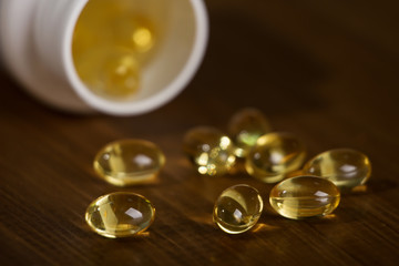 Capsules of fish oil spilled out open container on wooden table