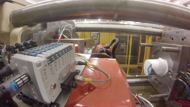 A wide-angle, point-of-view GoPro shot from a robotic arm in a factory, as it grabs a plastic product from an injection molding machine and carries it to another machine to apply a label. 
