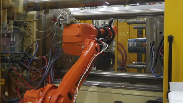 In a factory, a robotic arm grabs a plastic container from an injection molding machine and carries it to another machine to apply a label. 
