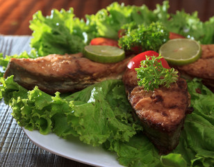 A piece of fried fish fillett served on blue glass table with lettuce, lime, tomato and herbs