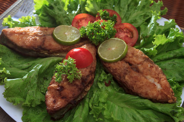 A piece of fried fish fillett served on blue glass table with lettuce, lime, tomato and herbs