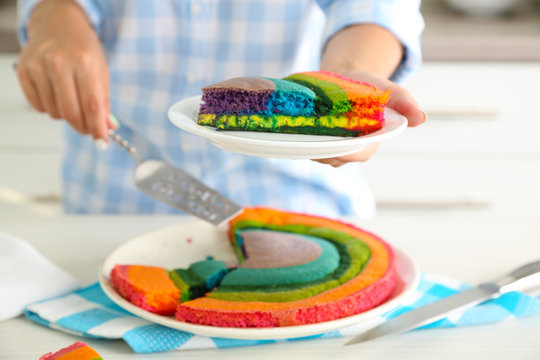 Young woman holding plate with portion of rainbow cake in kitchen