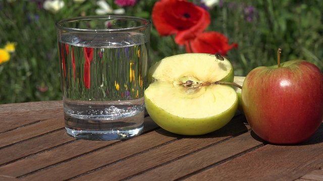 Glass Water and apple on table in the garden in front of summer flowers