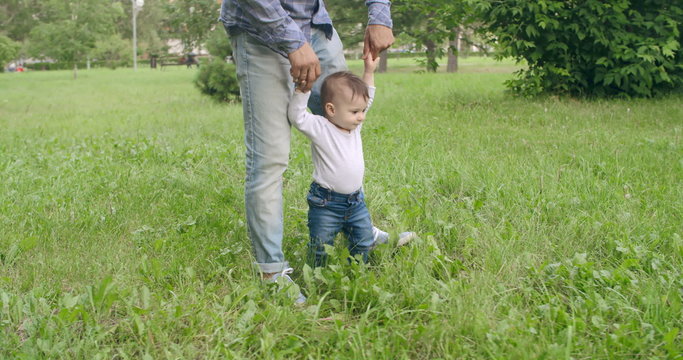 Adorable baby boy walking on grass helped by his dad 