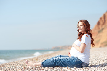 Fototapeta na wymiar Pregnant woman sitting at beach wearing casual clothes. Looking forward. Holding tummy. Motherhood. Maternity. 