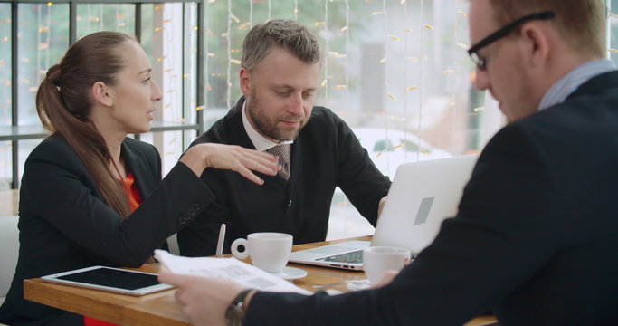 Businessman and businesswoman working on laptop at caf&eacute;, their colleague reading newspaper 