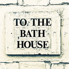 A weathered sign on a white door with the words 'To the Bath House'.