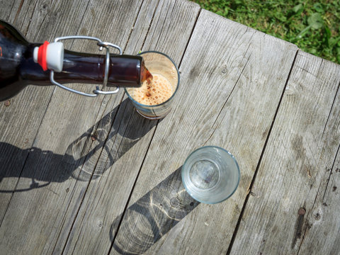 Frothy dark beer pouring into tall glasses from a brown glass bottle in summer garden on rustic wooden table