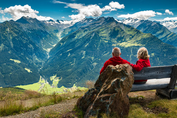 senior couple sitting in austrian alps and watching peaks with glacier and clouds - obrazy, fototapety, plakaty