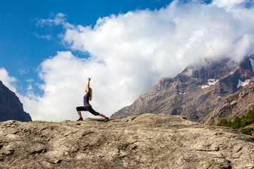 An attractive young woman doing a yoga pose for balance and stretching staying on top of high rock in the mountains sunny day blue sky with clouds