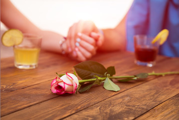 Obraz na płótnie Canvas Romantic scene flower Vintage natural wood cafe table beverage glasses rose flower on foreground lovers holding hands of each other young male female vacation style environment warm back-light sun