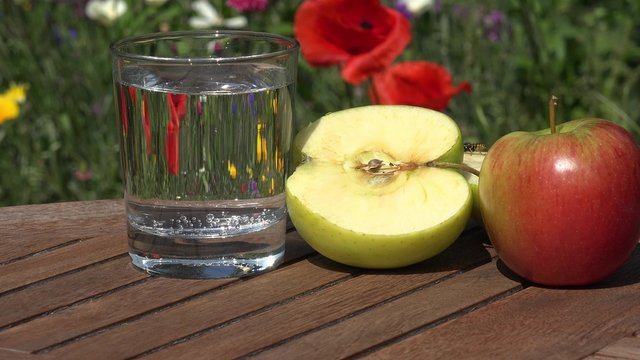 Glass Water and apple on table in the garden in front of summer flowers 