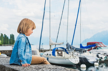Fototapeta premium Fashion portrait of a cute little blond boy sitting by the lake, wearing denim shirt and beige trousers