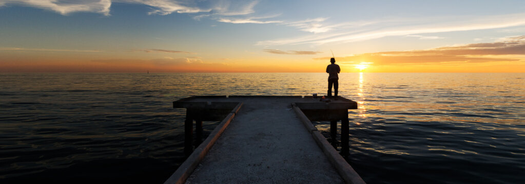 Panorama view of lonely man fishing alone during sunset at Bagan Sungai Burong Jetty with orange skies