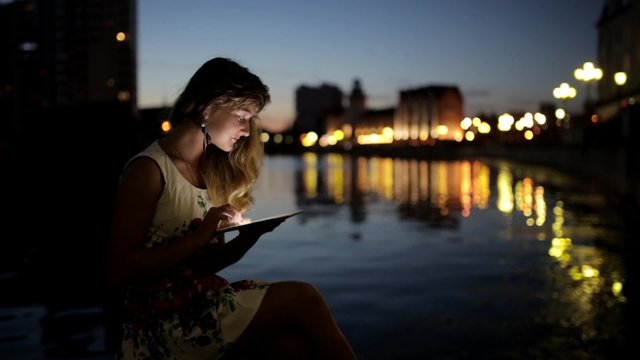 Teen girl using a tablet PC on the riverside of the city at evening time