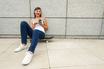 Fototapeta na wymiar Young girl siting on skateboard in the city with phone by the wa