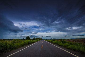 Asphalt road through the green field and clouds on dark sky in 