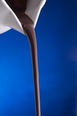 Chocolate Sauce Pouring From a White Pitcher Against a Blue Background