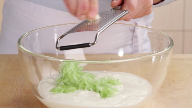 A garlic clove being grated into a cucumber and yogurt mixture
