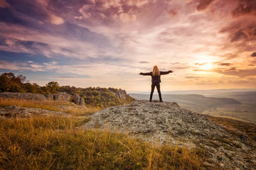 A woman on the top of a rock enjoys the view of sunset over an autumn forest