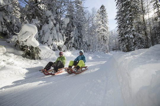 Germany, Bavaria, Inzell, couple having fun on sledges in snow-covered landscape