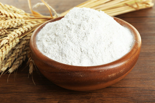 Whole flour in bowl with wheat ears on wooden table, closeup