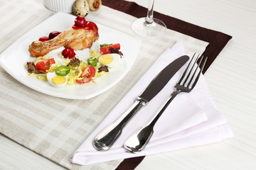 Dish of baked chicken leg and vegetable salad in white plate with glass of wine on table with napkin, closeup