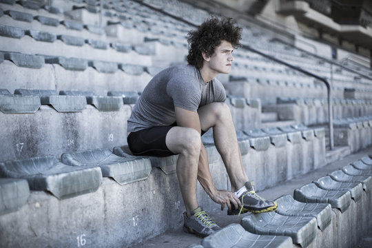 Young sportsman sitting on grandstand of a stadium tying his shoes
