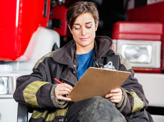 Fototapeta premium Firewoman Writing On Clipboard Against Trucks