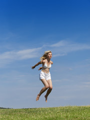 The happy woman in white bikini and shorts jumps in a summer green field against the blue sky..