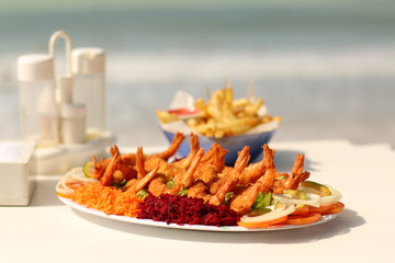 Giant shrimp served with beet and carrot salad and French fries with the blurred waves in the background
