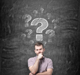 Brainstorming of young man. Question marks are drawn on the black chalkboard behind the person.