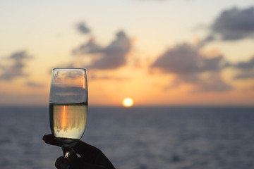 A glass of champagne raised up to toast at sunset on the ocean.