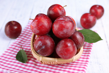 Ripe plums in wicker bowl on wooden table with napkin, closeup